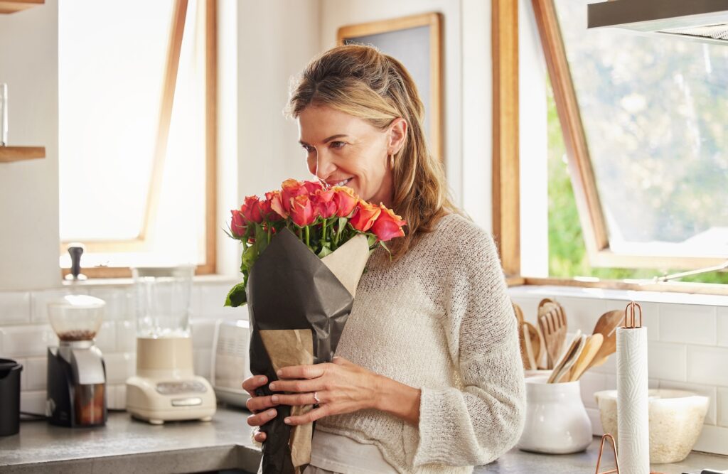 Flowers, smile, and elderly woman smelling rose in a kitchen, surprised by sweet gesture and or sec